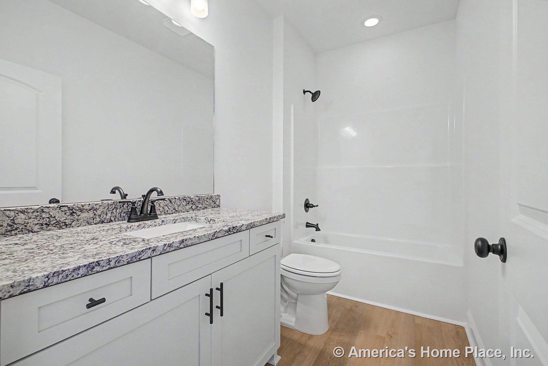 Granite vanity countertop with single sink, white shaker cabinets, matte black faucet and hardware, large wall mirror above, tub-shower unit with matching fixtures, wood-look