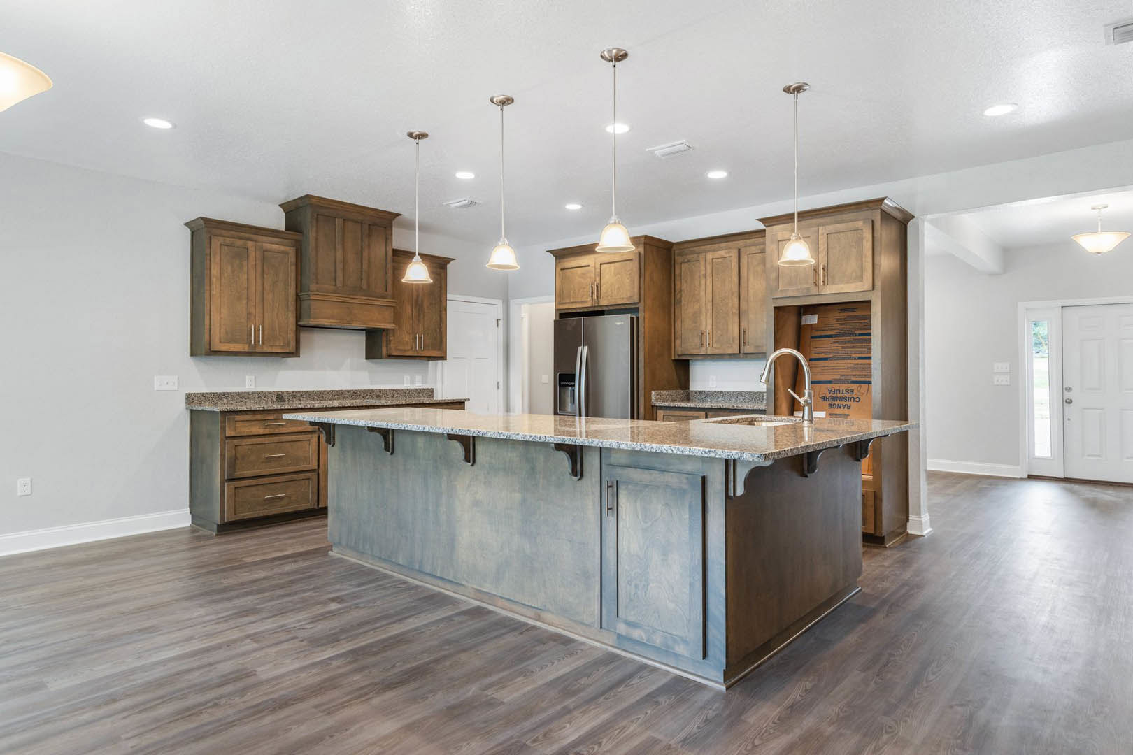 Spacious kitchen featuring a large granite island, white cabinetry, stainless steel refrigerator, modern ceiling light fixtures, and a white door with a silver handle.