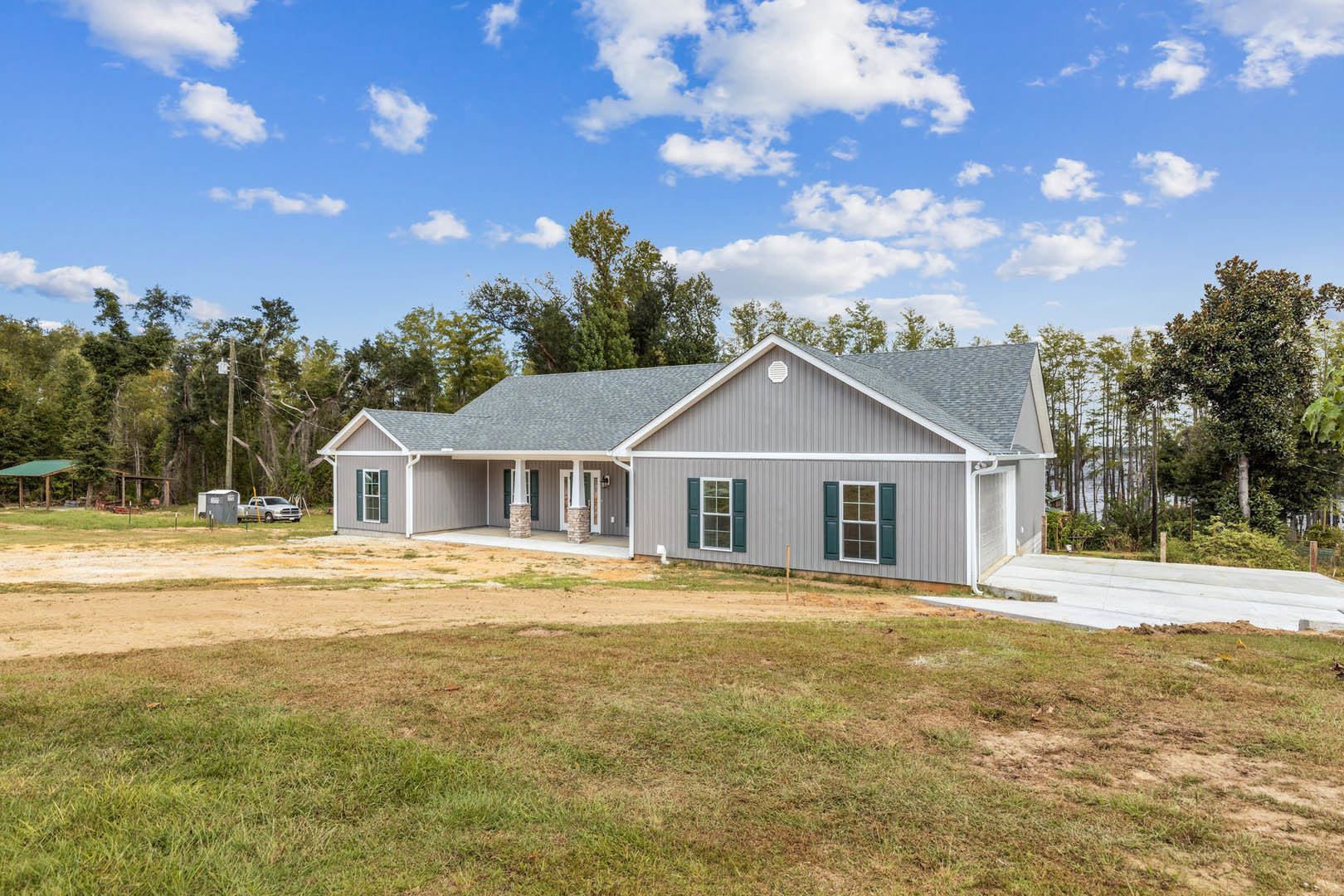 Two-story house with white exterior walls, green shuttered windows, front porch, driveway leading to parked car, grassy lawn bordered by a fence, trees and blue sky with scattered