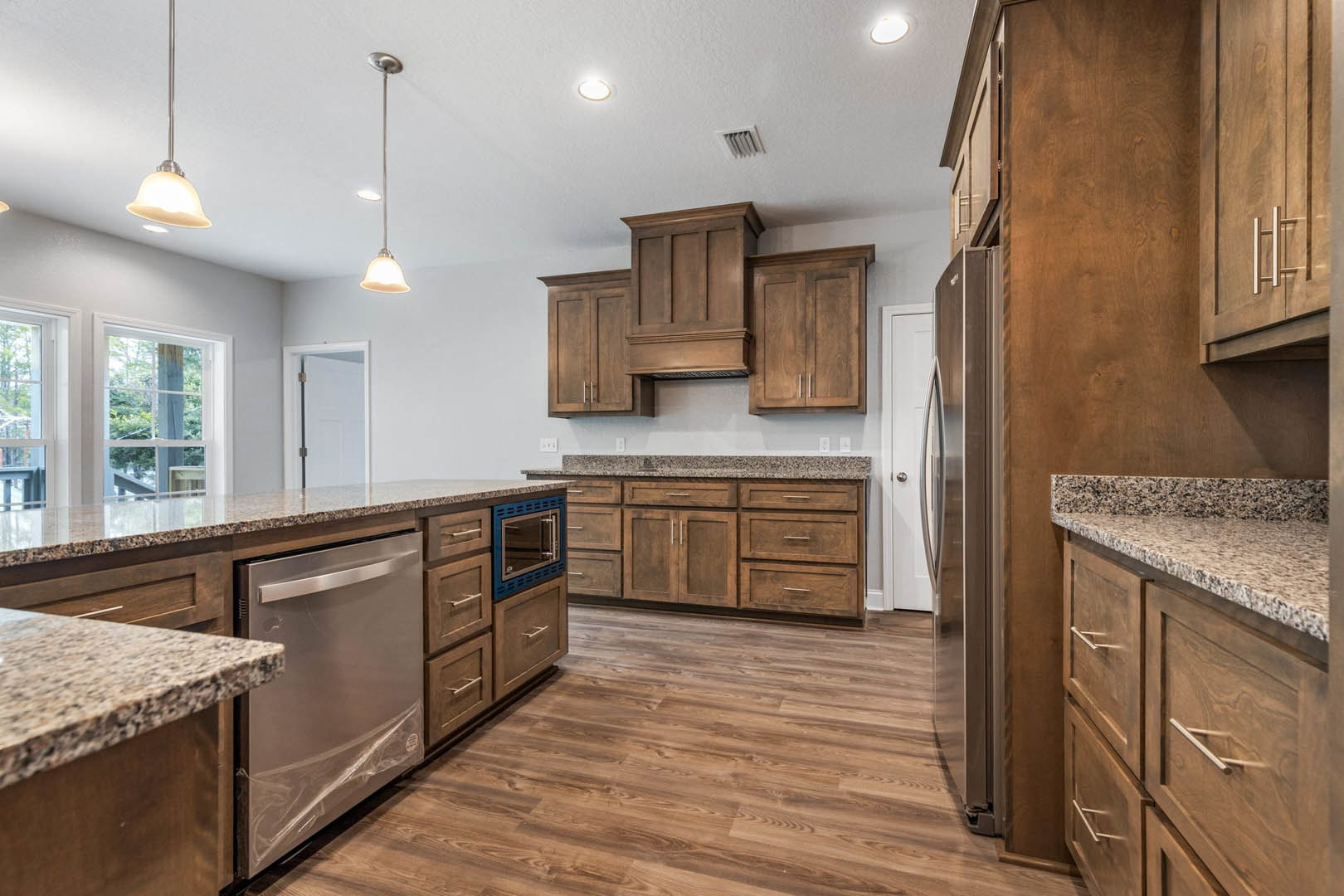Kitchen with wood plank flooring, brown granite countertop, white cabinetry, stainless steel refrigerator, and a white door with metal hinge.
