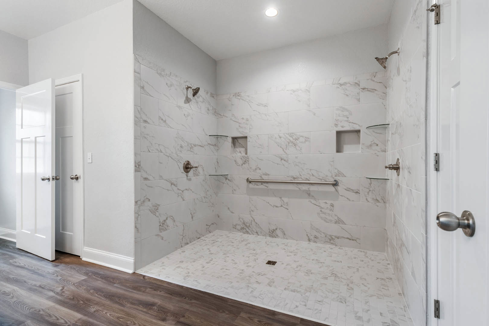 Bathroom featuring marble tile walls, wood floor, ceiling light, white marble shower with metal bar, white tile floor with drain, and close-up views of white doors
