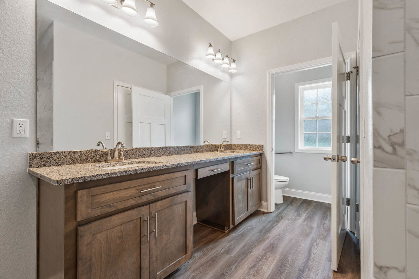 Bathroom with marble countertop, wood floor, white electrical outlet on wall, modern light fixture, toilet, window, sink and chrome faucet on counter, white cabinetry and drawers.