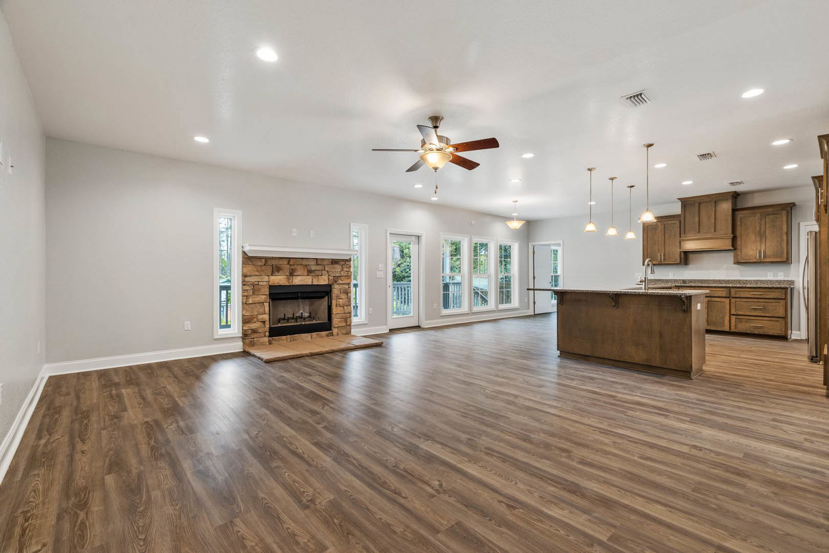 Spacious living room featuring hardwood floors, central fireplace with black frame, ceiling fan with light, marble-topped brown countertop, and large window overlooking trees