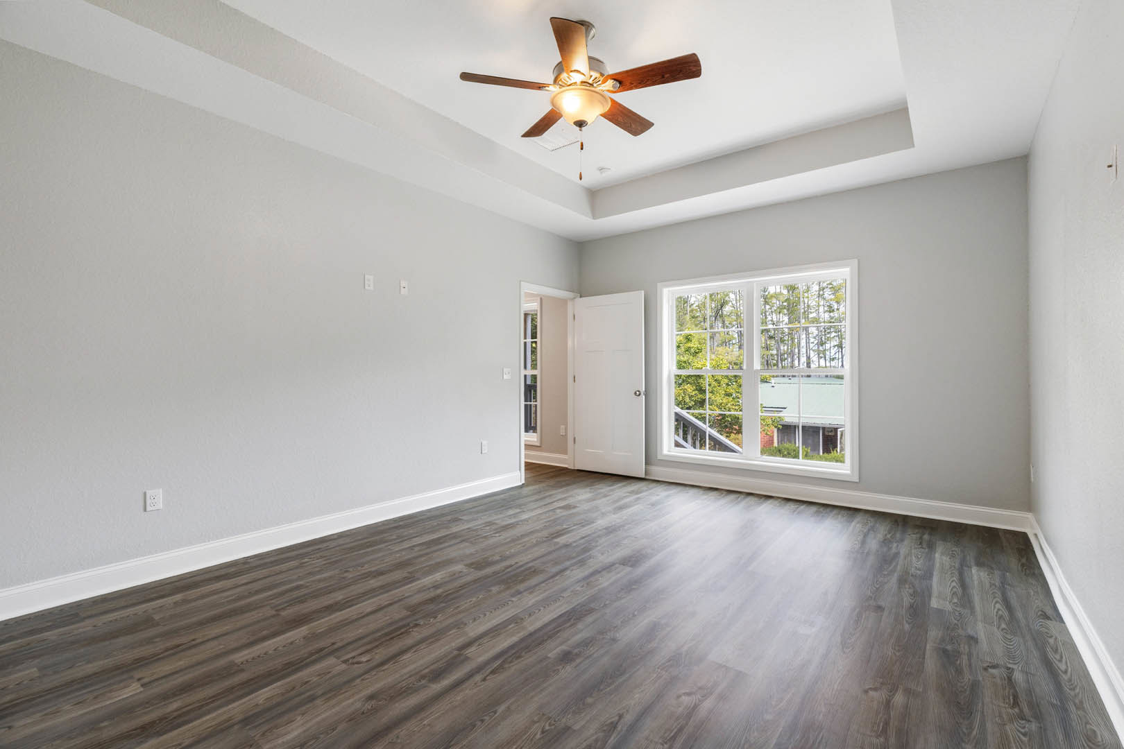 Ceiling fan with light fixture, wood flooring, white walls, window showing trees outside, white door with silver knobs