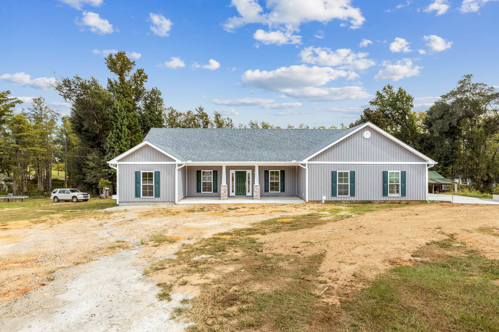 Two-story house with green door and glass panels, green shuttered windows, dirt yard, white car parked beside dirt road, surrounded by mature trees under cloudy sky