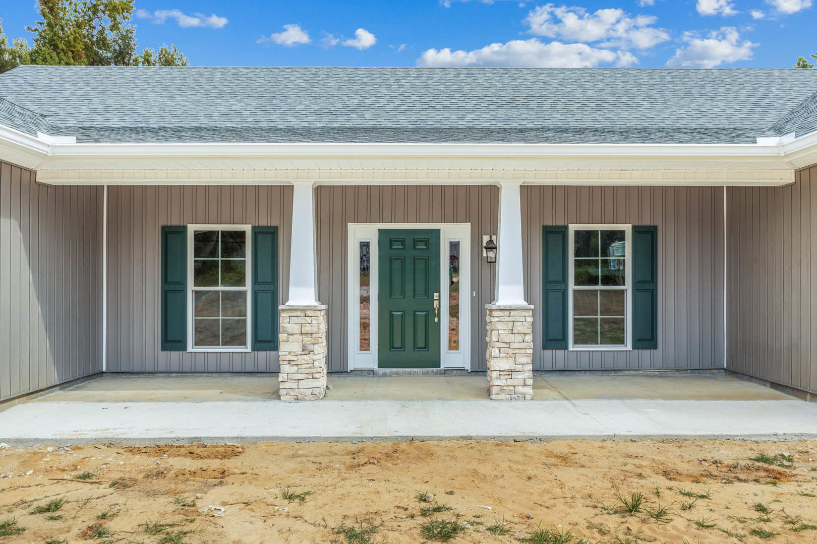 Green front door with white frame, white column on porch, light-colored siding, gabled roof, landscaped dirt area in foreground, large windows, cloudy sky above