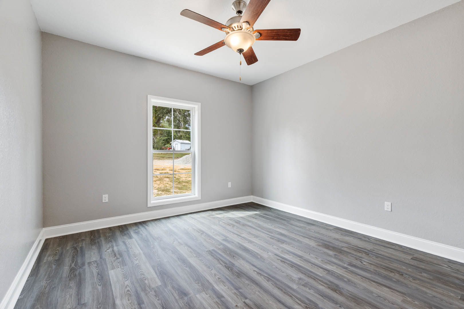Ceiling fan with integrated light fixture mounted on white plaster ceiling, wood flooring, large window with view of grassy field and white building, neutral walls with crown