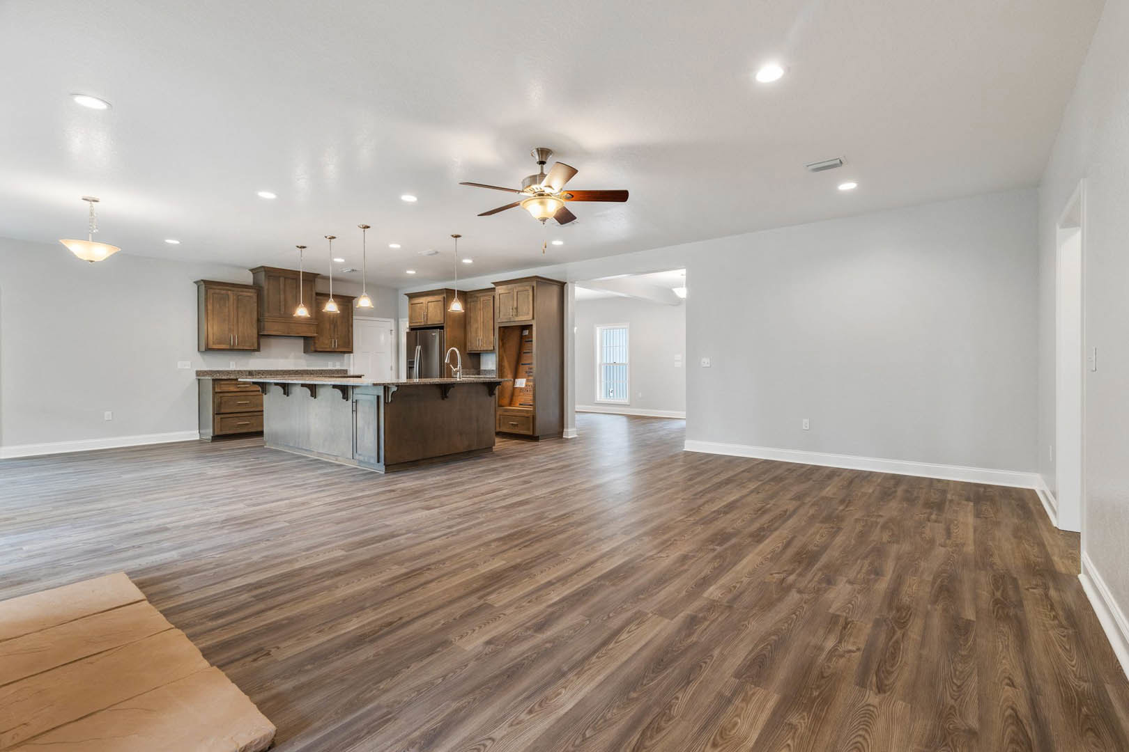 Open kitchen and living room with hardwood floors, white cabinetry, ceiling fan with lights, bright recessed lighting, window with white frame, and a piece of paper on the floor.