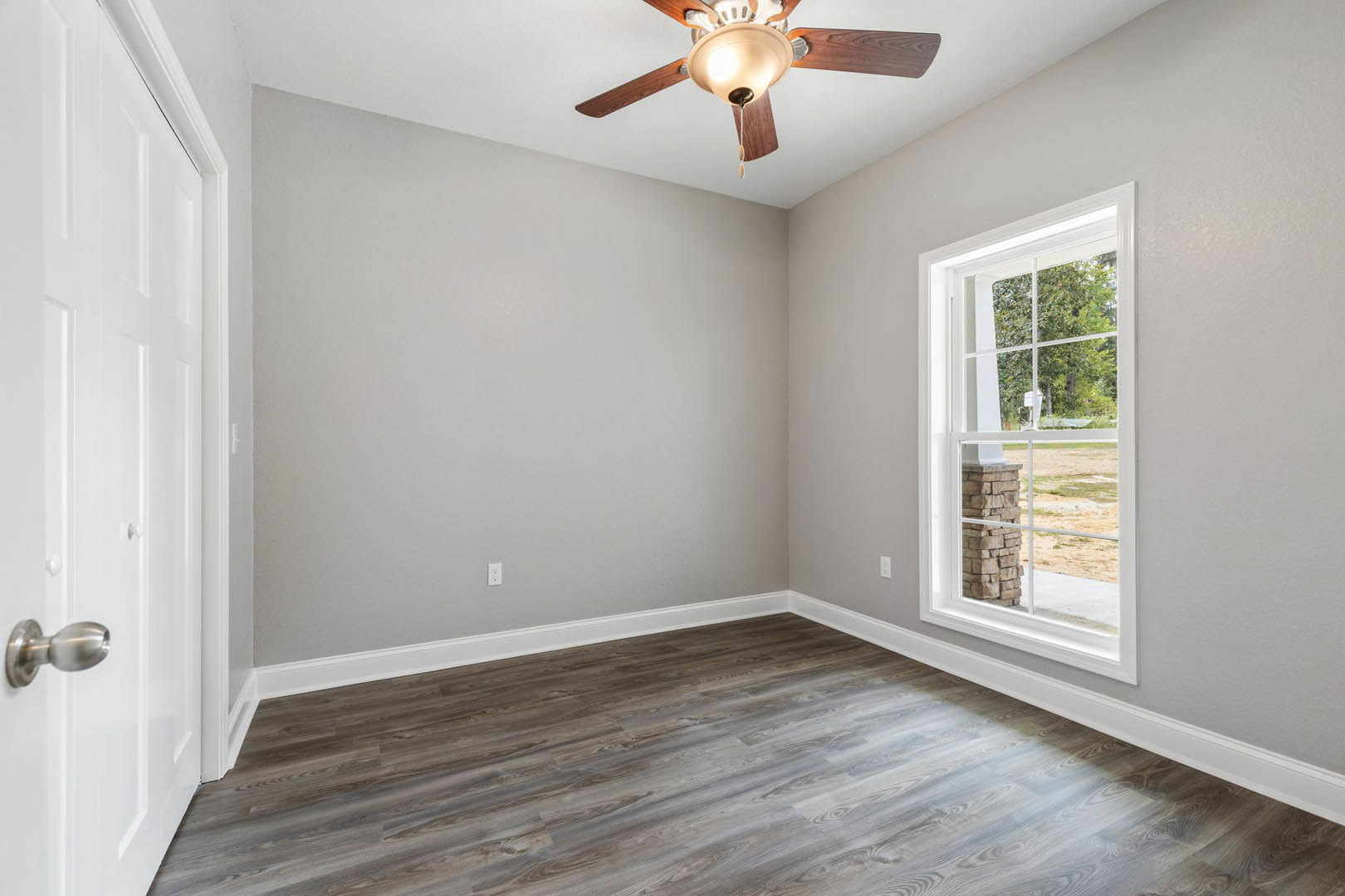 Ceiling fan with light fixture above wood flooring, large window showing tree and grass outside, white walls with molding and plaster finish