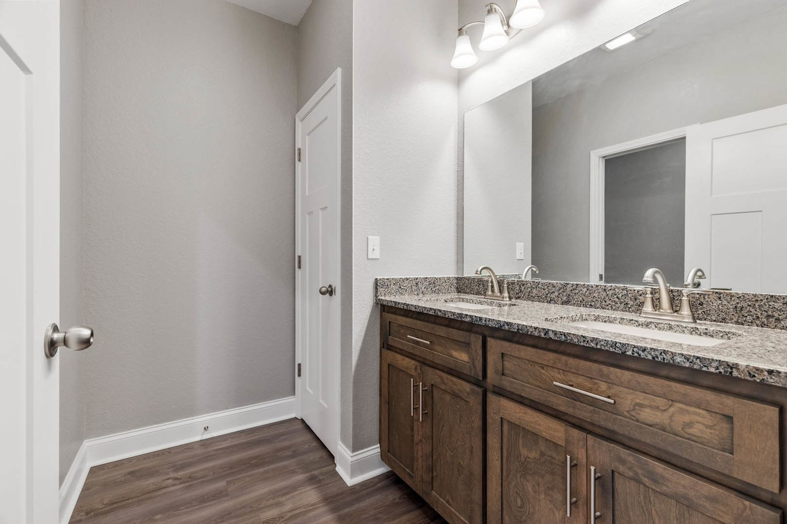 Bathroom with granite countertop vanity, rectangular mirror, chrome faucet, white door with silver handle, wood flooring and white baseboard, tile backsplash, window with natural
