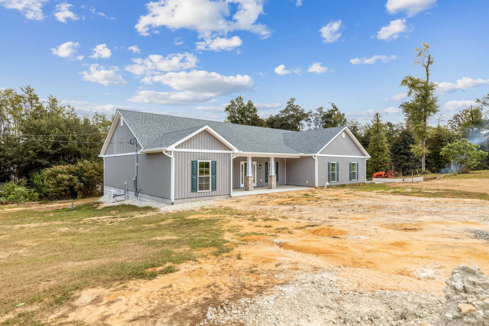 Two-story home with white-framed windows, covered front porch, expansive dirt yard bordered by mature trees, under a partly cloudy blue sky
