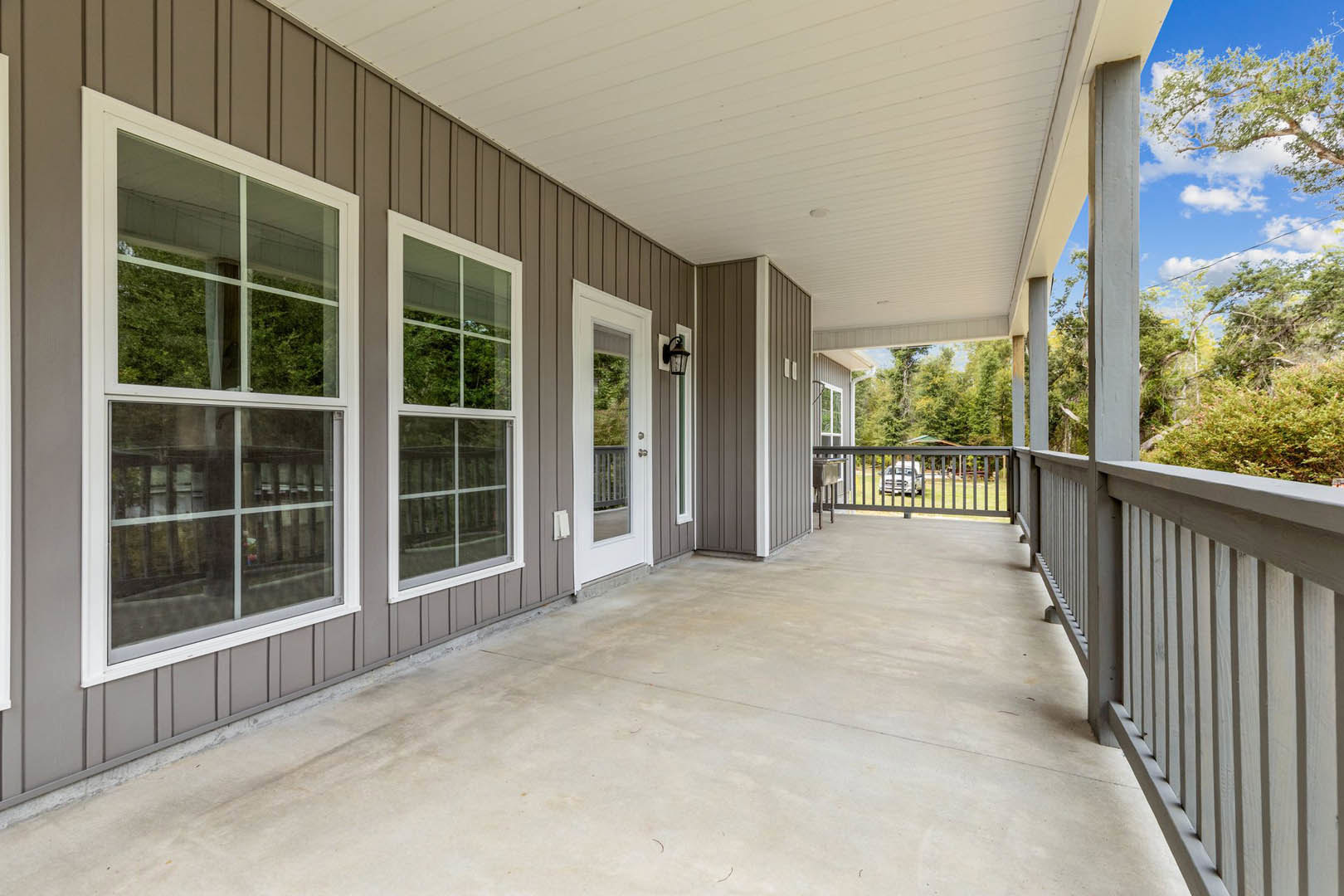Two porches with railings and windows, white framed windows, white door with glass panel, concrete patio, car behind a fence, trees and sky in background