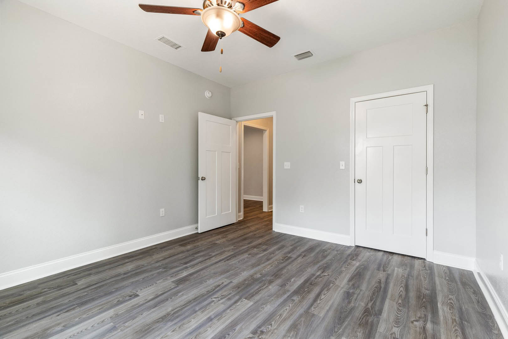 Wood flooring, white paneled doors with silver knobs, ceiling fan with light fixture, smooth white walls and ceiling