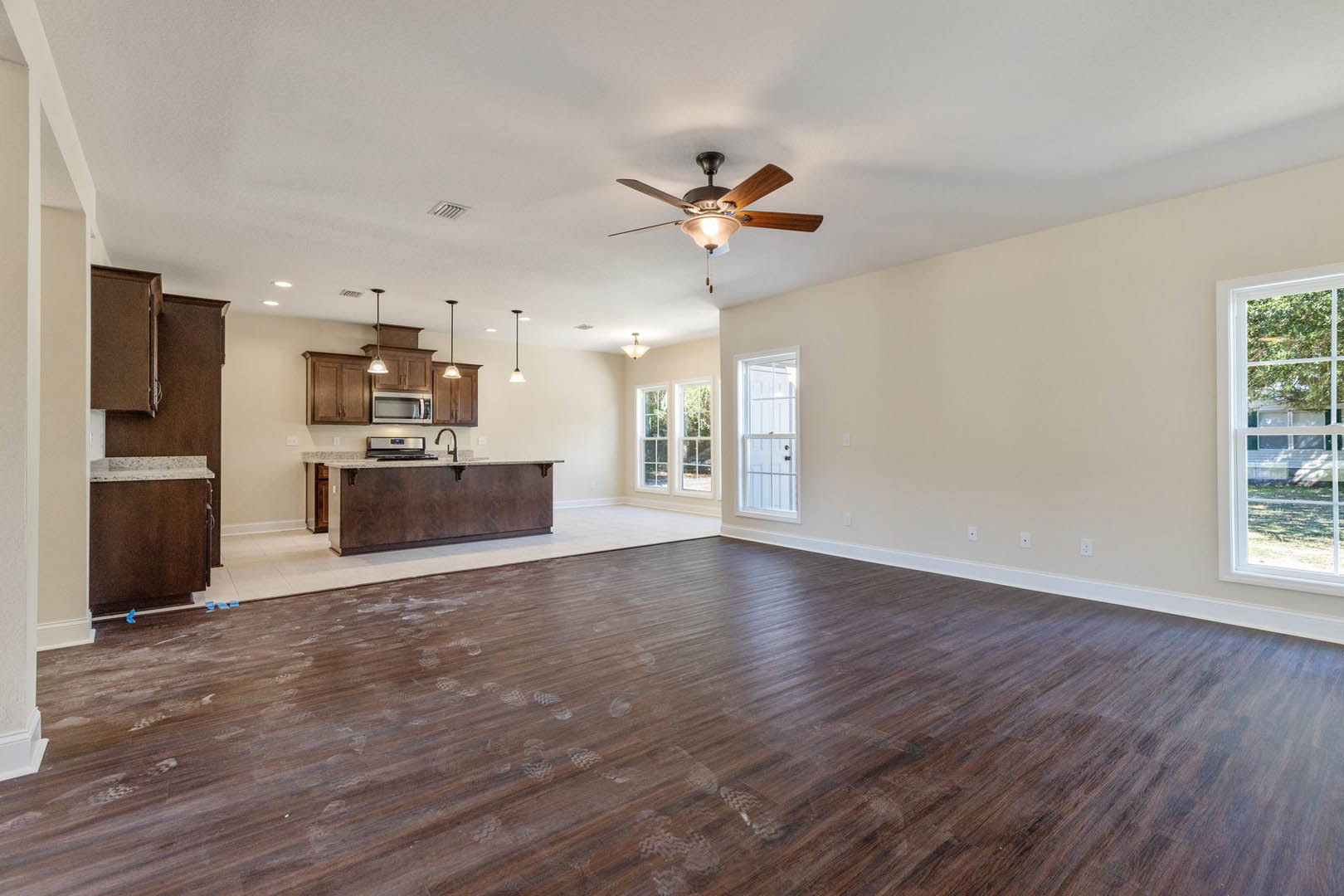 Spacious open room featuring wood flooring with visible footprints, modern kitchen with white cabinetry and stone countertops, ceiling fan with light fixture, white-framed window