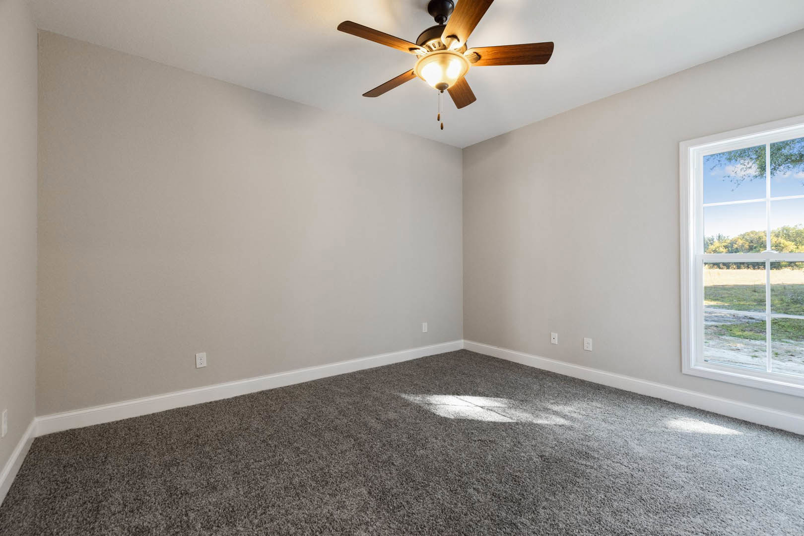 Carpeted bedroom with white walls, ceiling fan with light fixture, large window overlooking grassy field, crown molding along ceiling edges