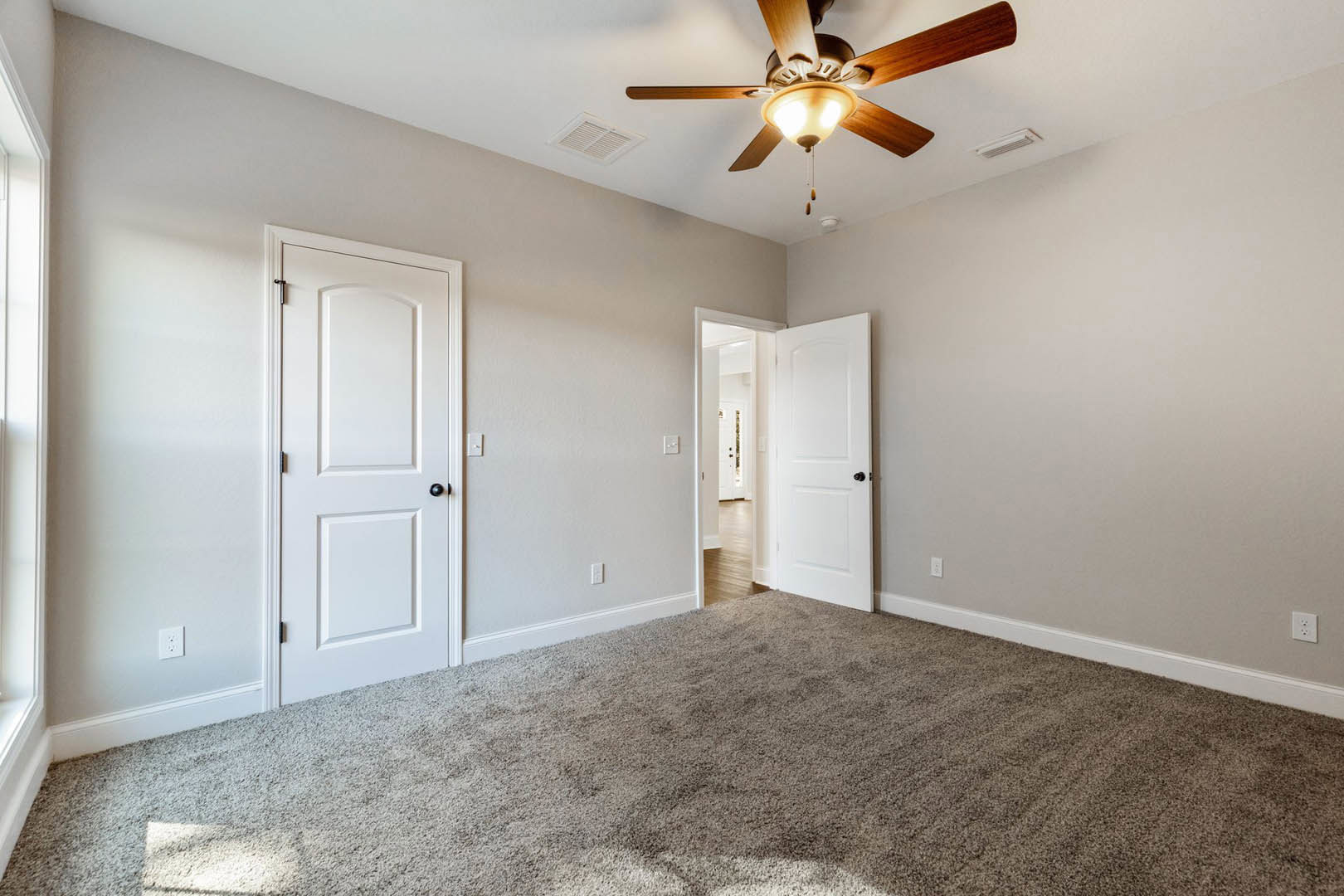 Carpeted room with white walls, ceiling fan with light, two white doors featuring black knobs, and ceiling vent