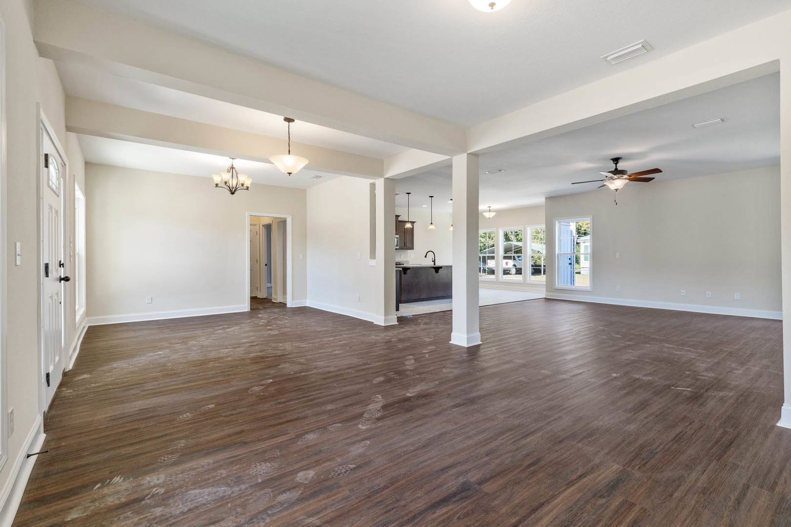 Spacious empty room with polished hardwood flooring, white plaster walls, ceiling fan with light, chandelier, and door illuminated by overhead fixture