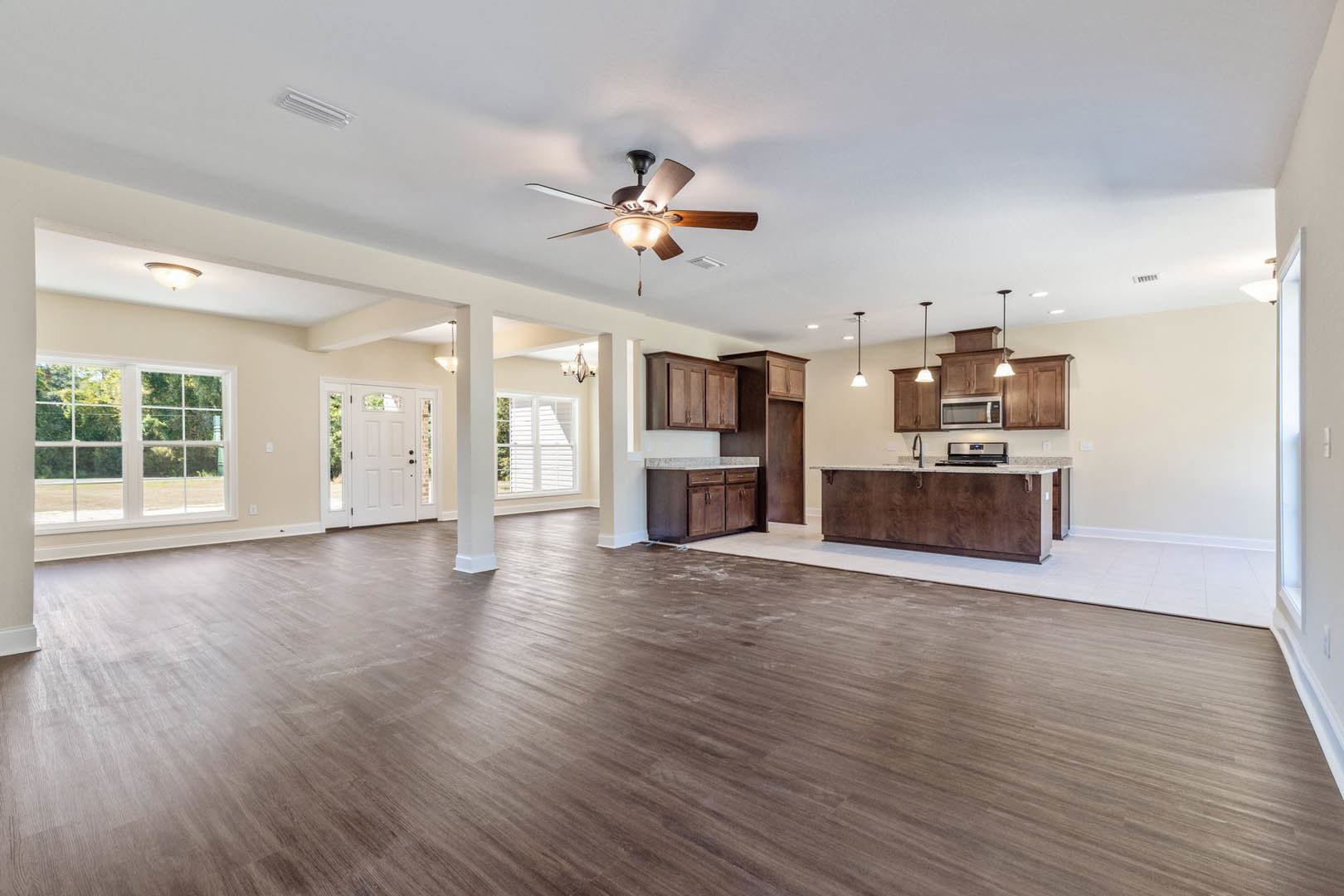 Spacious open floor plan featuring hardwood flooring, kitchen with white countertops and brown wooden cabinetry, dining area, ceiling fan with light, white door with window, and