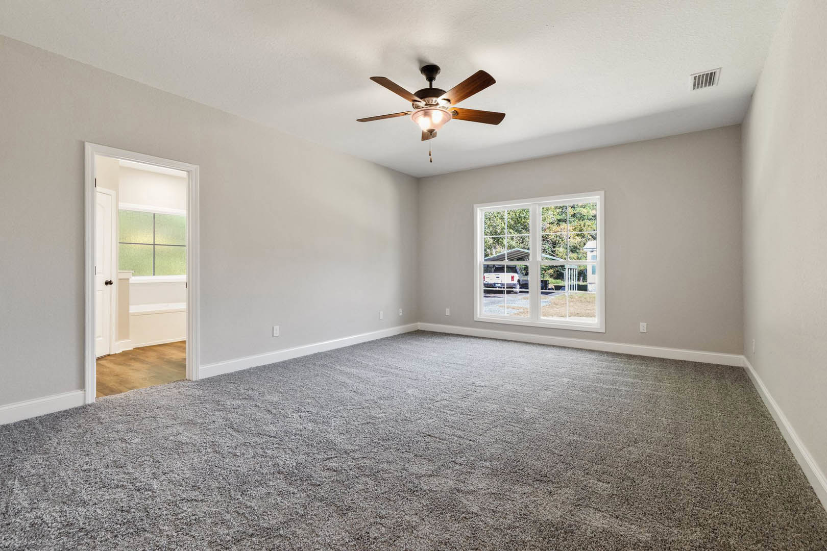 Ceiling fan with light fixture, grey carpet flooring, white-framed window, open door to adjacent room, wall vent, neutral painted walls