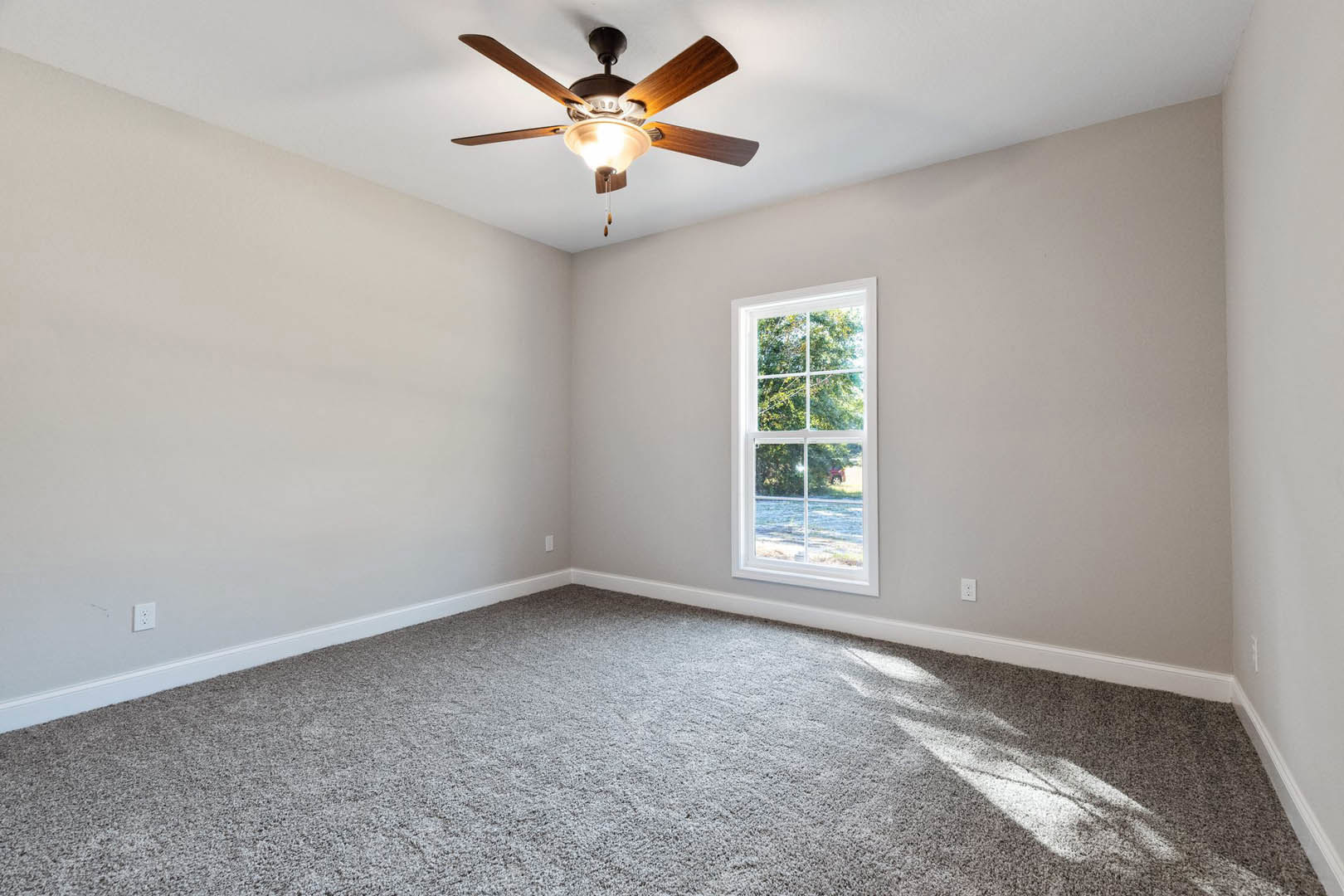 Bedroom with beige carpet, white walls, ceiling fan with light fixture, large window overlooking trees and lawn, crown molding along ceiling.