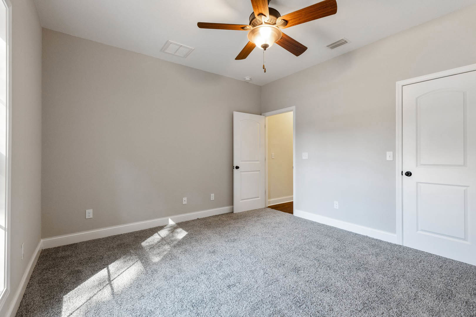 Carpeted room with white walls, ceiling fan with light, white door featuring black handle, and square ceiling vent