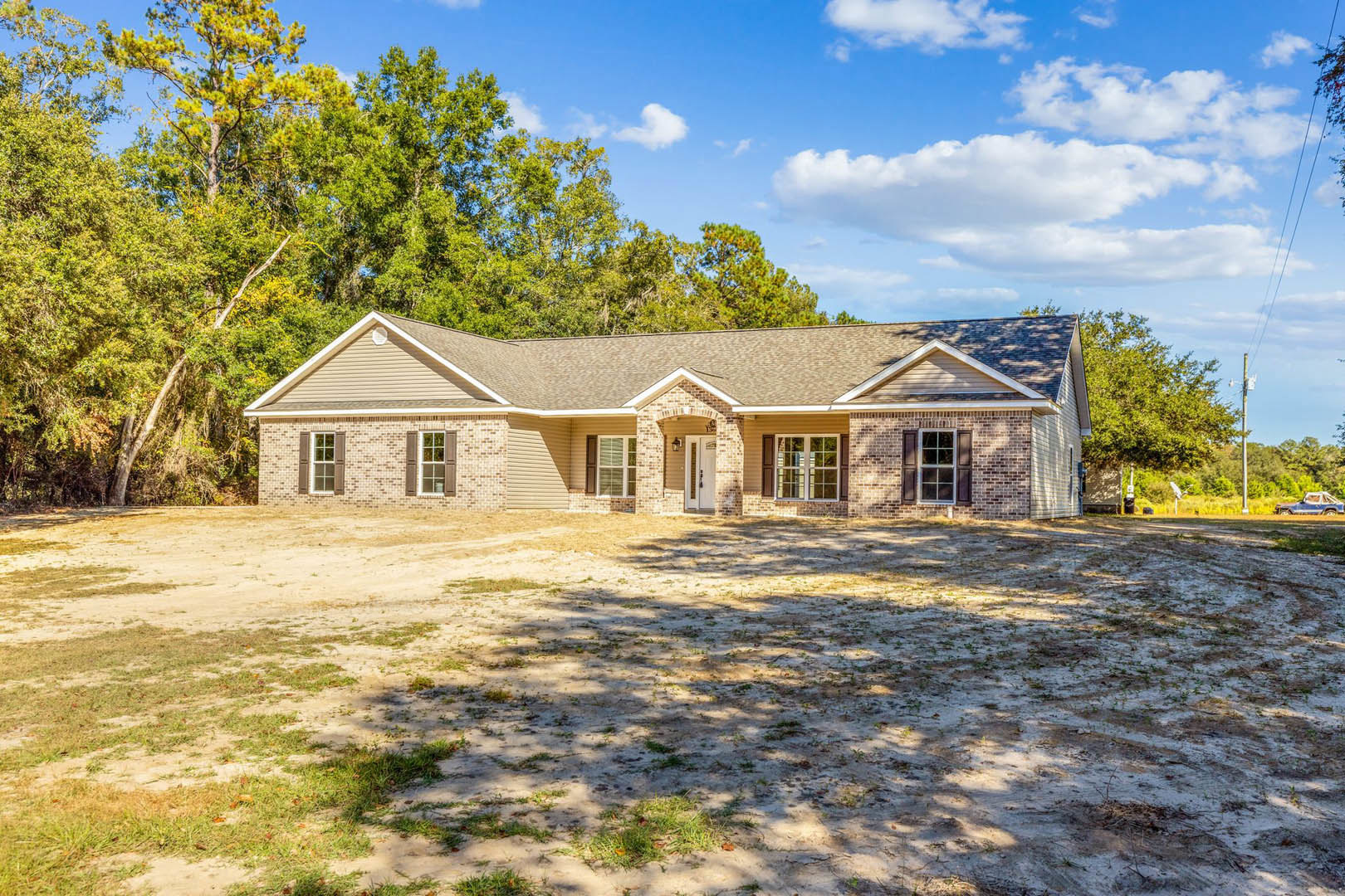 Single-story house with gray siding, white front door, white-framed windows with closed blinds, and a dirt yard in front.