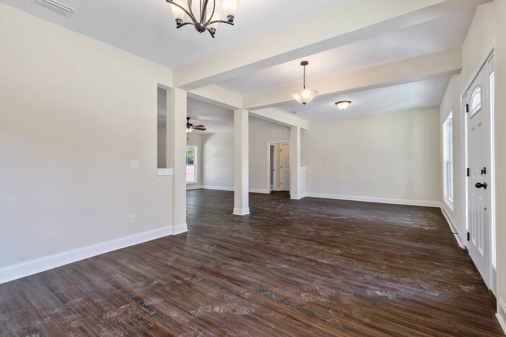 Spacious room with polished hardwood floor, white pillars, ceiling fan, chandelier, and white door illuminated by wall light