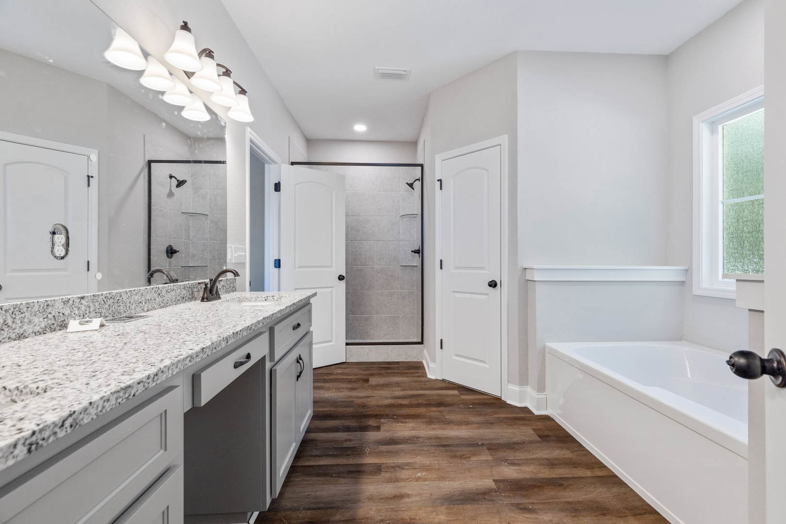 Bathroom featuring a freestanding bathtub, white sink with black hardware, white paneled door with black knobs, tile flooring, white-framed window, row of wall-mounted lights, and