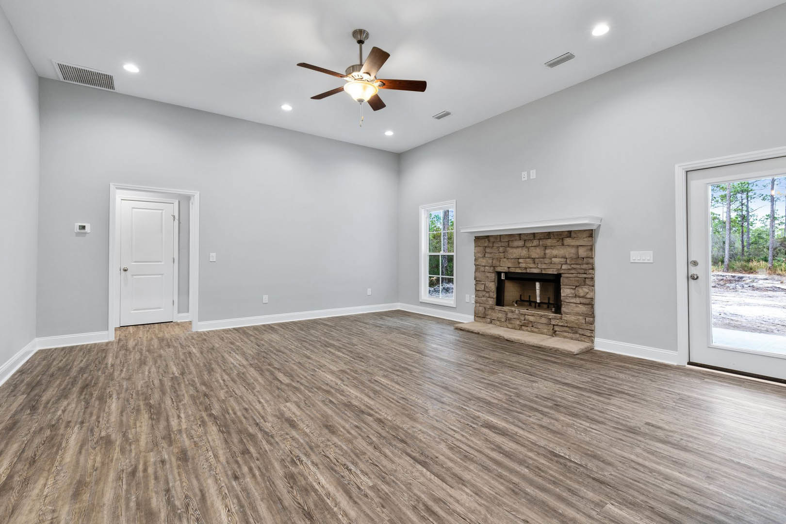 Living room with hardwood flooring, stone fireplace, ceiling fan with light, white door with silver handle, window showing tree outside