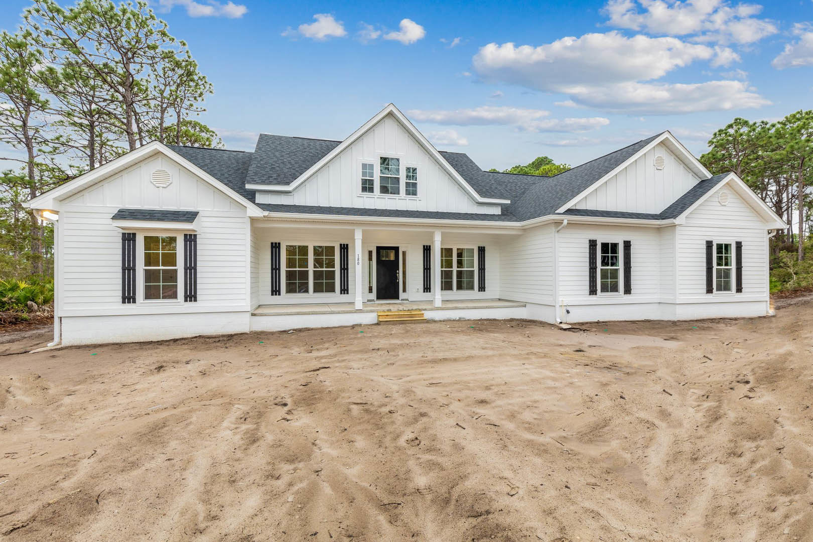 White house with black trim and black door, glass-paned windows, sandy dirt lot in front, steps leading to entry, cloudy sky overhead.
