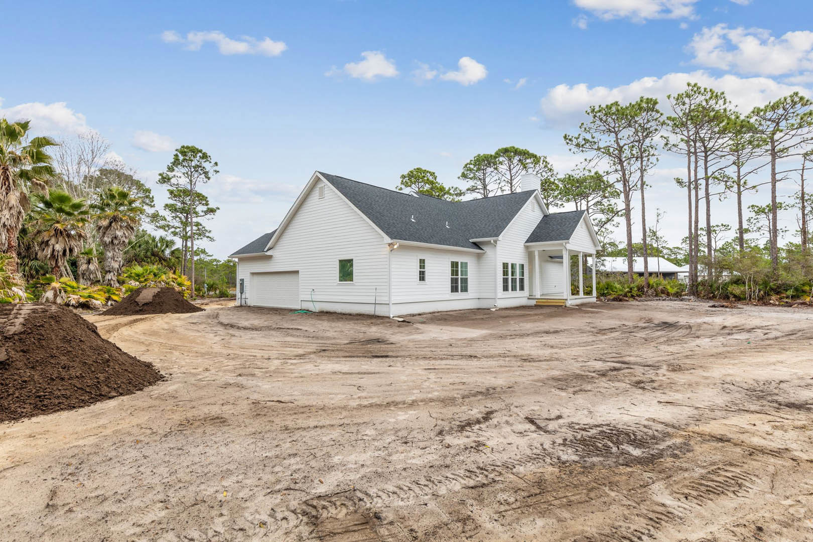 Partially built house with exposed framing, surrounded by piles of dirt and tall green trees under a cloudy sky