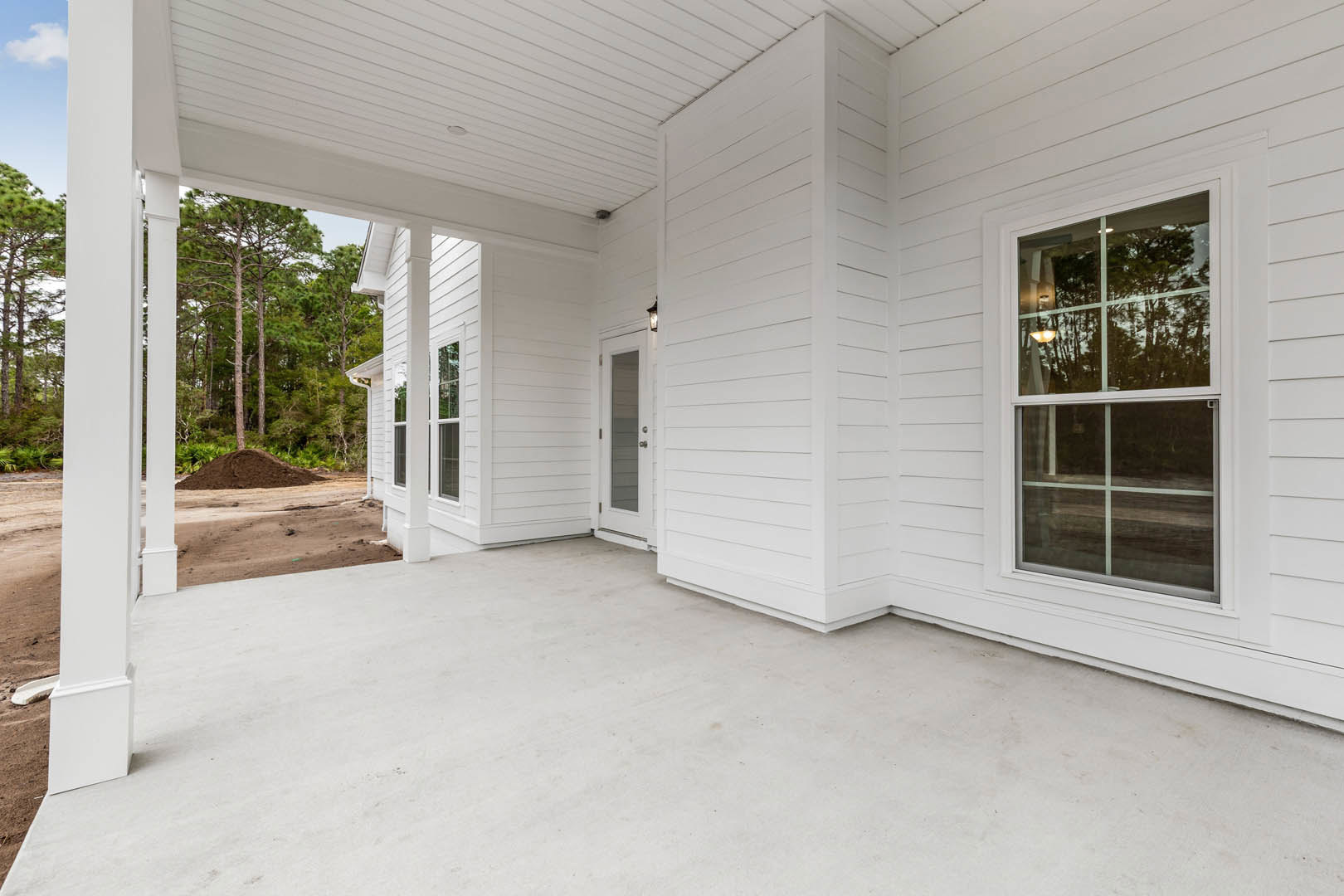 White exterior home with covered porch, glass panel door, illuminated window, white concrete floor, wood plank ceiling, and surrounding trees.