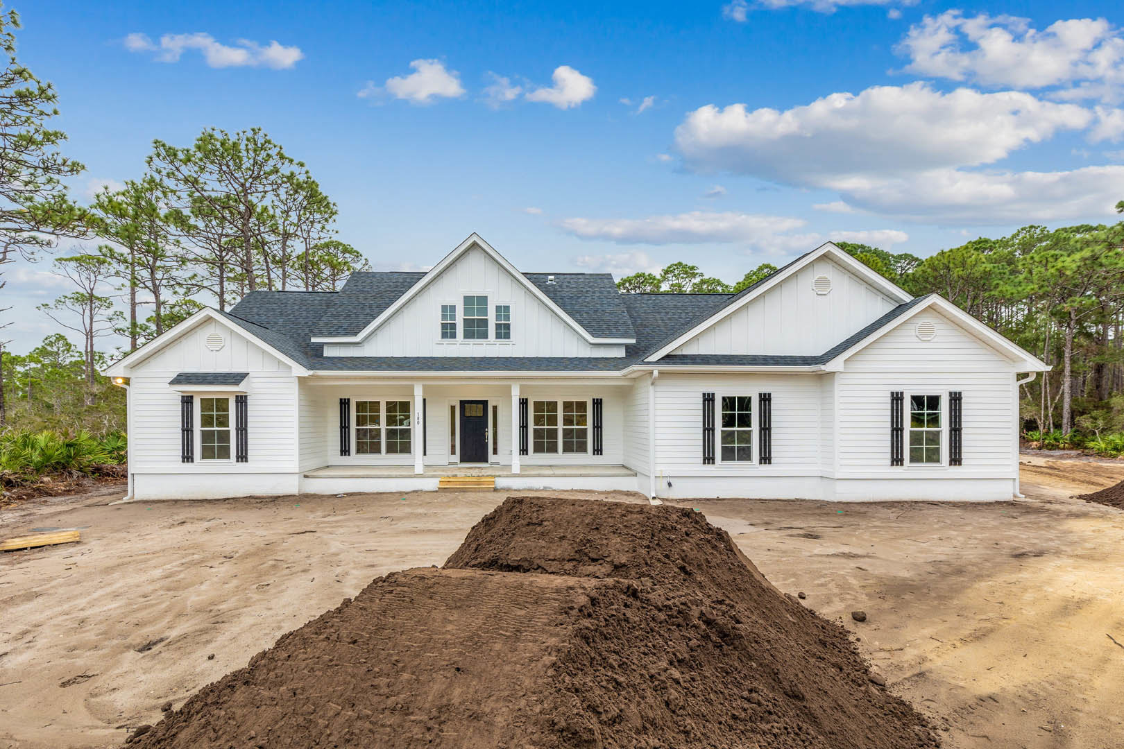 White house with black door and multi-pane windows, large mound of dirt covering front yard, cloudy sky overhead.