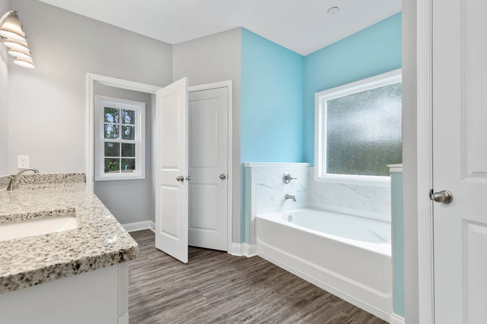 White freestanding bathtub beside a modern sink with chrome faucet, light tile flooring, frosted glass window showing trees, and minimalist countertop with stacked white plates.