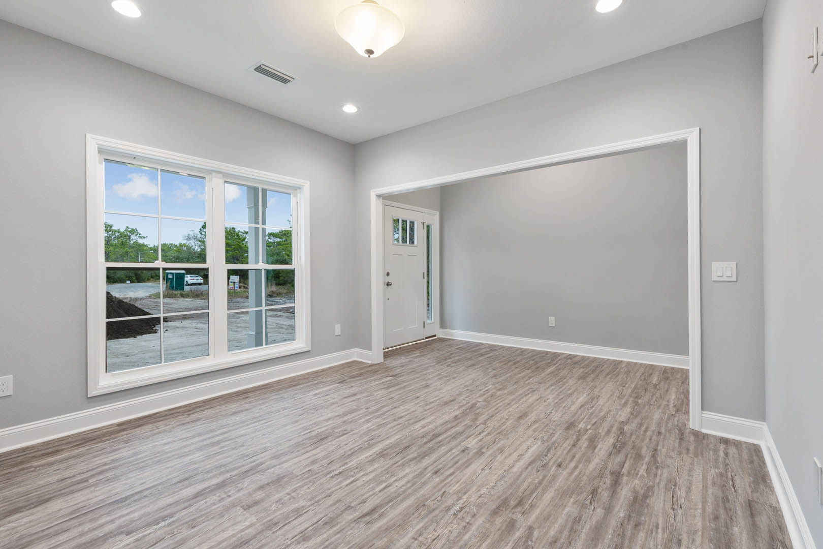 Room with wood flooring, white walls, large window overlooking construction dirt, white door with glass panel, close-up of modern light fixture, white light switch on wall.