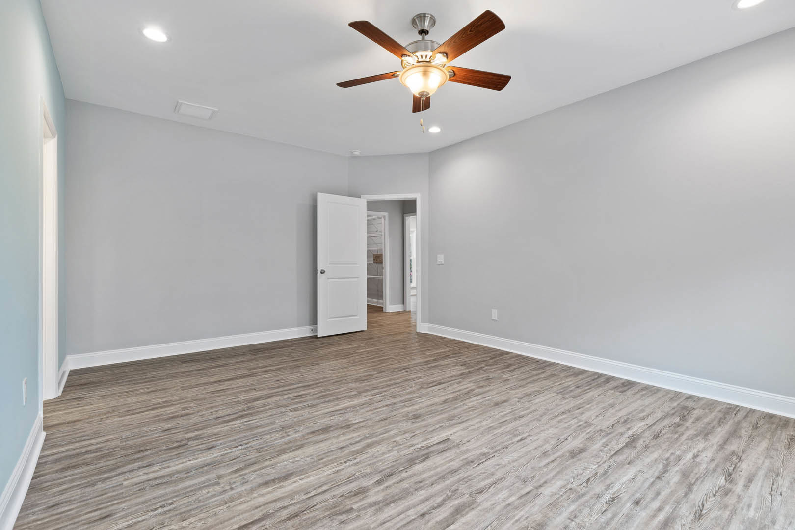 Room with wood laminate flooring, white plaster walls, ceiling fan with integrated light fixture, and white door with silver knob opening into adjacent space
