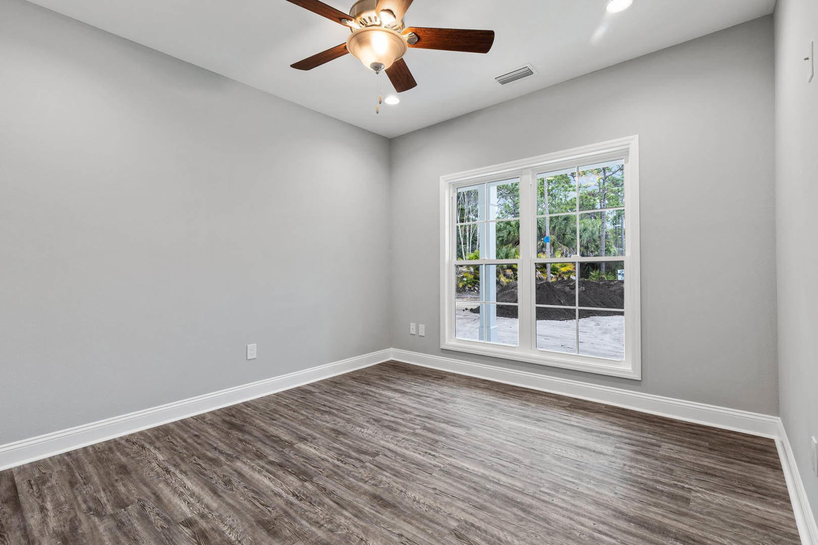 Wood flooring in a room with white walls, ceiling fan with light, window overlooking dirt path and trees, visible vent and molding along baseboard