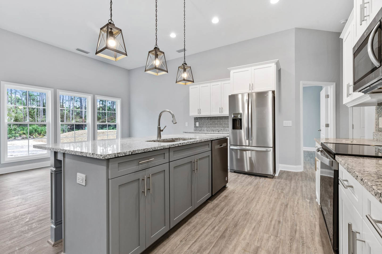 Spacious kitchen featuring a large central island with quartz countertop, stainless steel refrigerator, white cabinetry, modern pendant lights, and a window overlooking leafy trees