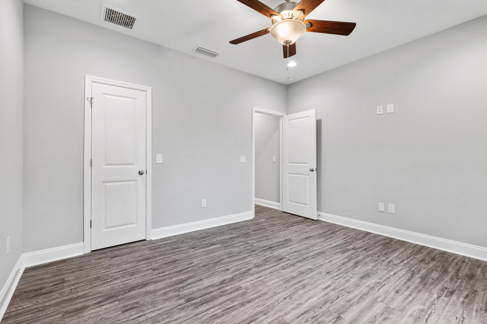 Wood flooring and white walls in a room with a ceiling fan and light fixture, two white doors with silver handles, and a white vent on the wall.