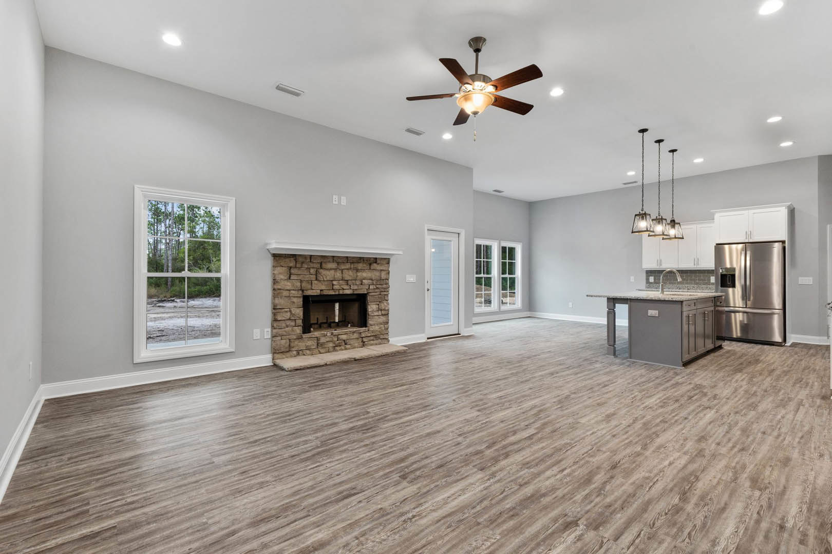 Spacious open floor plan featuring hardwood floors, stone fireplace, ceiling fan with light, stainless steel double-door refrigerator, and large window overlooking forest