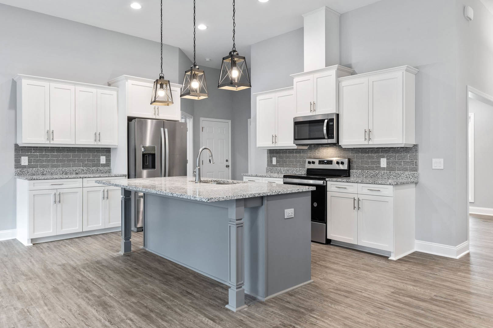 Modern kitchen featuring a central island with undermount sink, stainless steel refrigerator, granite countertops, white cabinetry, built-in microwave, and pendant light fixture.