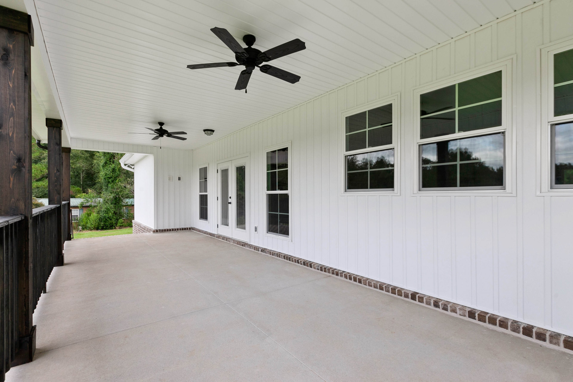 White exterior home with black ceiling fan featuring wooden blades on covered porch, concrete flooring, large windows reflecting nearby trees, and close-up view of window trim.