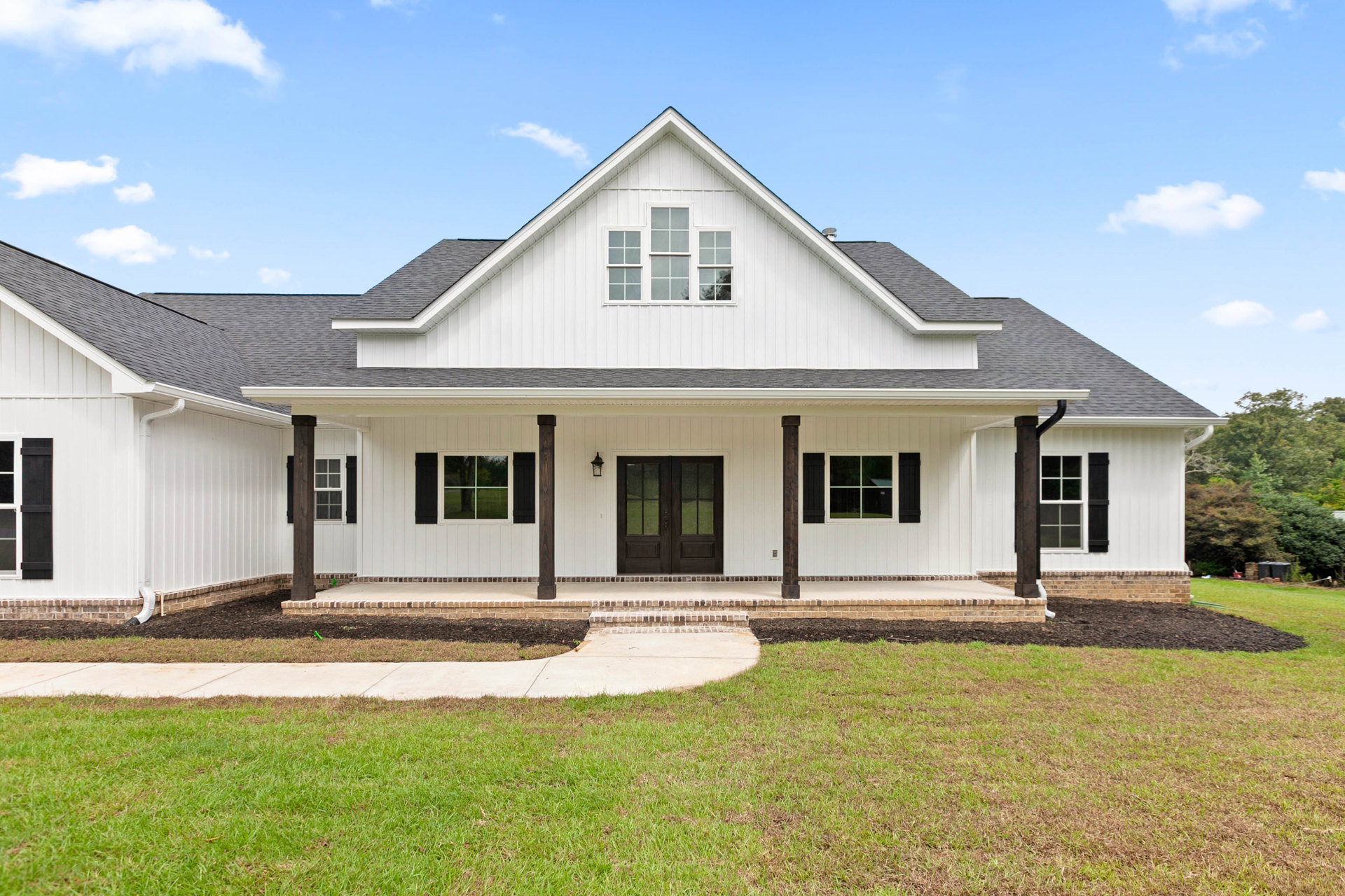 White siding house with black-trimmed windows, double glass-paneled front doors, brick and concrete walkway, manicured lawn, tree visible through window, partly cloudy sky overhead