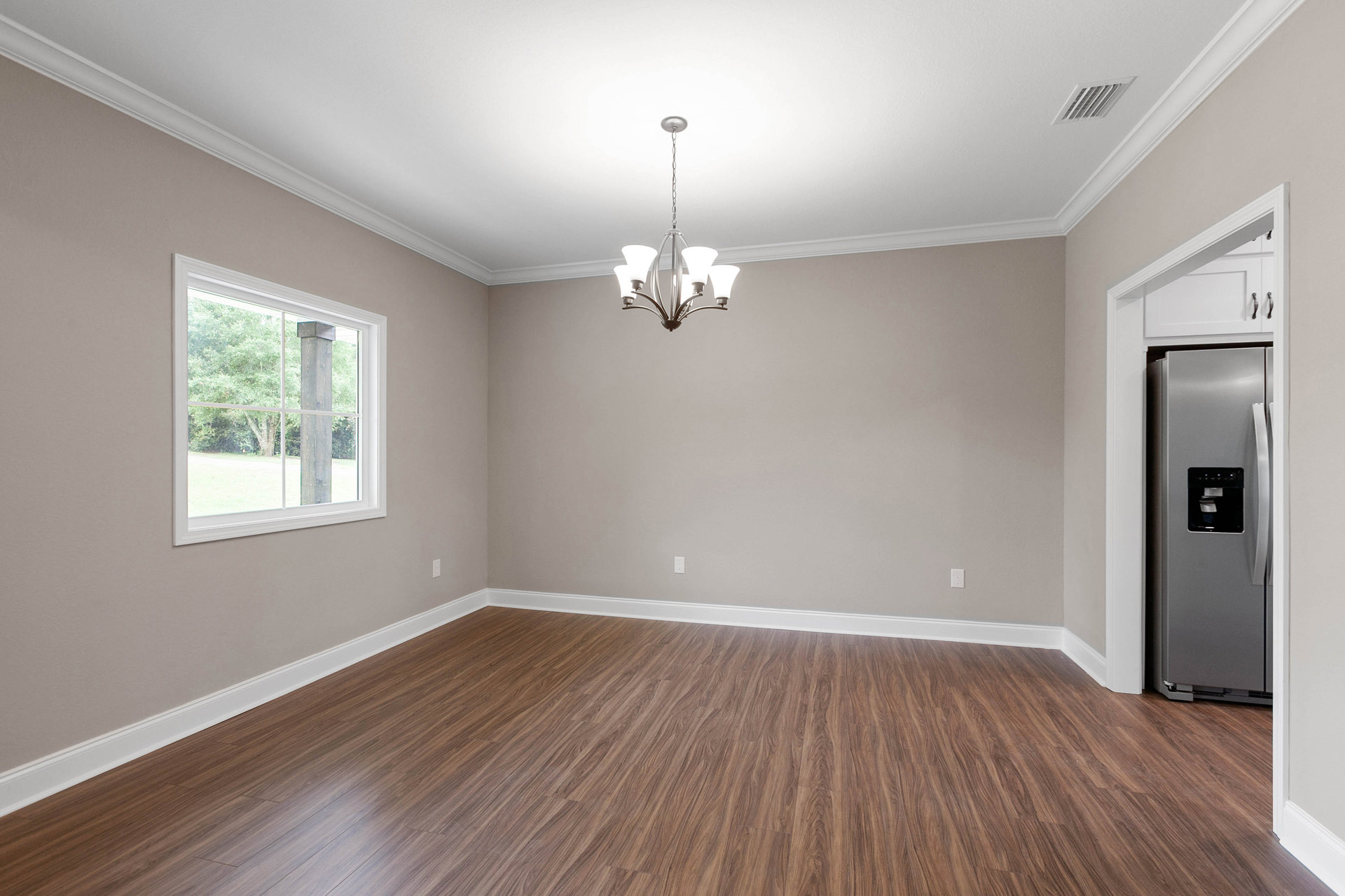 Open living space with wood flooring, white baseboards, modern chandelier, refrigerator partially visible, large window framed by exterior wood post, ceiling vent overhead