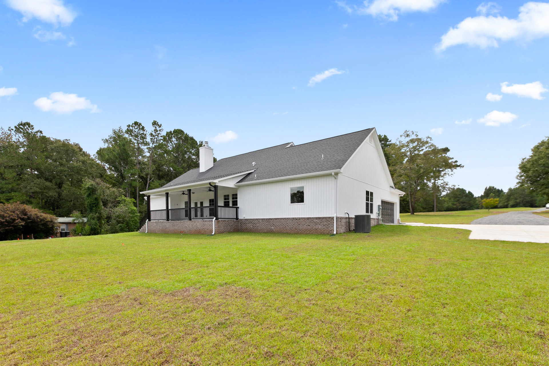 White farmhouse with black railings, expansive green lawn bordered by a brick wall, leafy trees in the background, and a black air conditioner unit beside the house