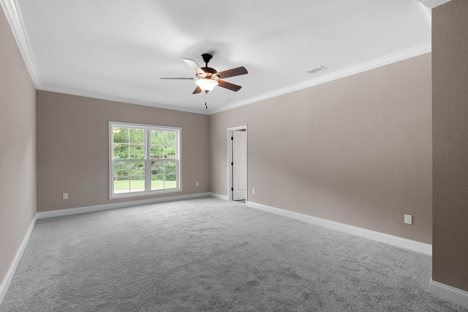 Bedroom with light gray walls, ceiling fan with integrated light, white trim, open white door with black handle, large window overlooking green trees, hardwood flooring