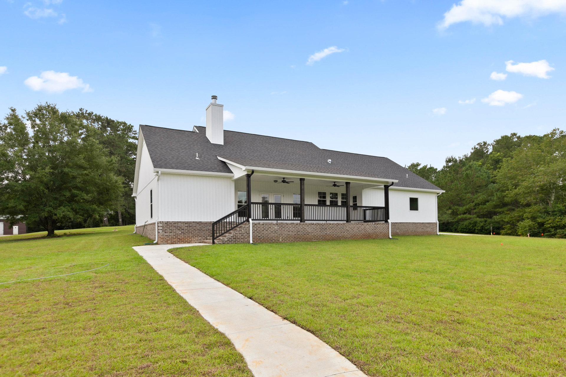 Brick house with covered porch, concrete walkway leading to entrance, grassy lawn, mature tree, white trim, gabled roof with chimney, partly cloudy sky