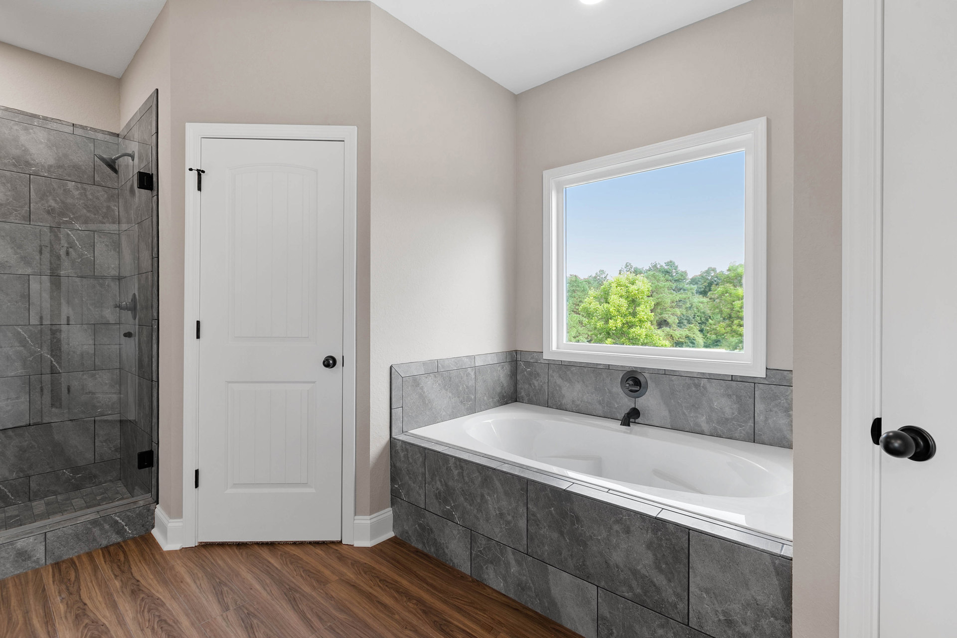 Modern bathroom with freestanding white bathtub beneath large window overlooking trees, glass shower enclosure, white door with black hardware, light tile flooring, chrome faucet
