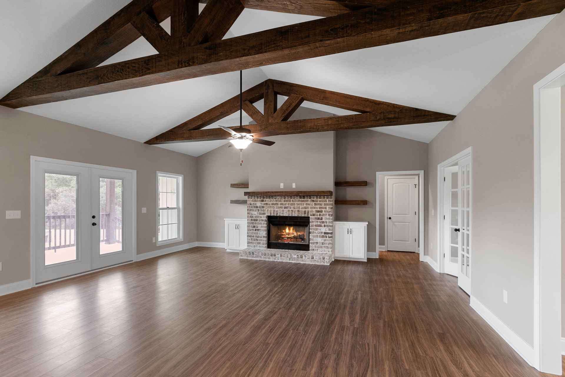 Living room with hardwood floors, stone fireplace with fire, exposed wooden ceiling beams, white double doors with glass panes, white door with black handle, large window with trim