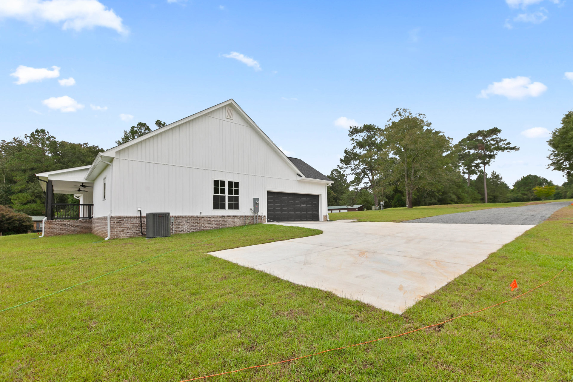 White house with brick accent wall, concrete driveway bordered by grass and trees, attached garage, large grey HVAC unit, and close-up window detail.